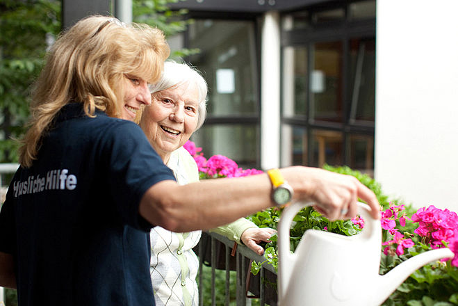 Das Bild zeigt eine Mitarbeiterin des BRK, die einer älteren Dame beim Blumen gießen hilft.