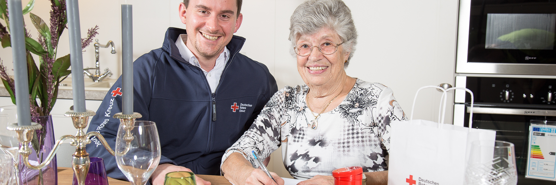 Das Foto zeigt Rotkreuzmitarbeiter mit älteren Dame in deren häuslicher Umgebung mit Rotkreuzdose