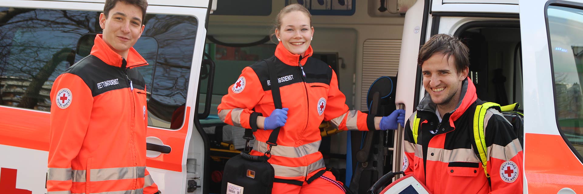 Foto zeigt junge Frau und zwei junge Männer in Rettungsdienstkleidung am Einsatzwagen