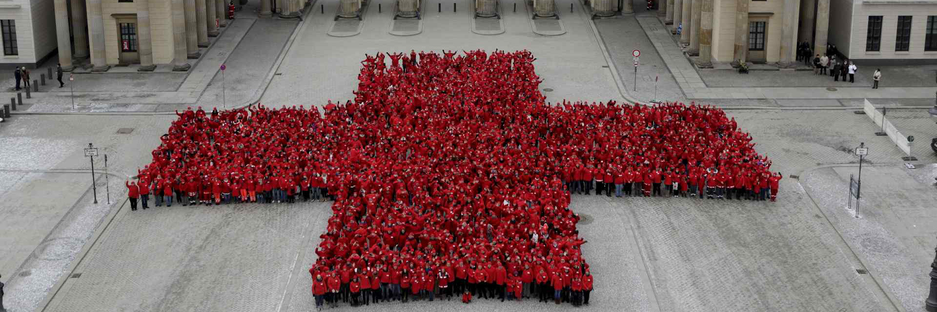 Bild zeigt Mitarbeiterinnen und Mitarbeiter des Roten Kreuzes. Sie bilden gemeinsam die Form eines Kreuzes vor dem Brandenburger Tor in Berlin.