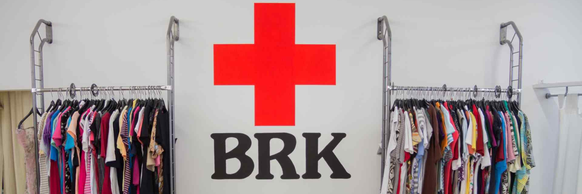 Das Bild zeigt das BRK Logo. Links und rechts befinden sich Kleiderstangen mit Kleidung.
