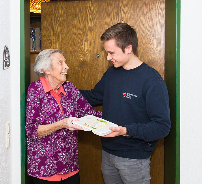 Bild zeigt Freiwilligen bei älterer Dame und bringt ihr ein Mittagsmenü